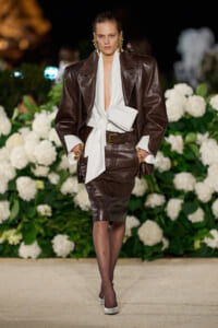 Model walking a runway in a dark brown leather suit with a white blouse tied at the waist; floral backdrop behind her.