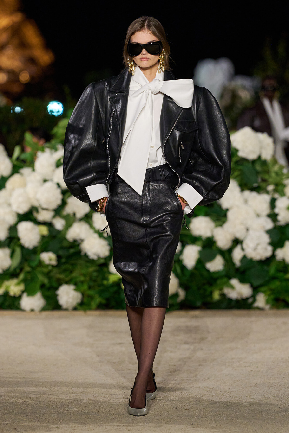 Model on a runway wearing an oversized black leather jacket, white blouse with a large bow, and a black leather pencil skirt, with sunglasses and gold earrings, standing against a backdrop of white flowers.