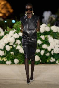 Runway model in an all-black leather ensemble: sleeveless vest and knee-length skirt, oversized sunglasses, gold earrings and bracelets, white heels, nighttime backdrop of white flowers.
