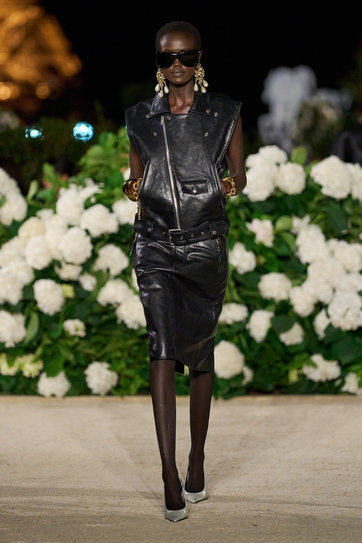 Runway model in an all-black leather ensemble: sleeveless vest and knee-length skirt, oversized sunglasses, gold earrings and bracelets, white heels, nighttime backdrop of white flowers.