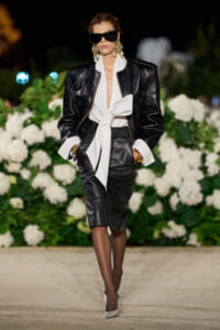 Model walking the runway in a black leather suit with a white bow-blouse, oversized sunglasses, and gold earrings, against a backdrop of white florals.