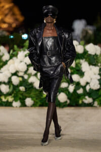 Model in a black leather ensemble (jacket, corset-style top, belt) with a cap and sunglasses walking a runway, white hydrangea backdrop behind.