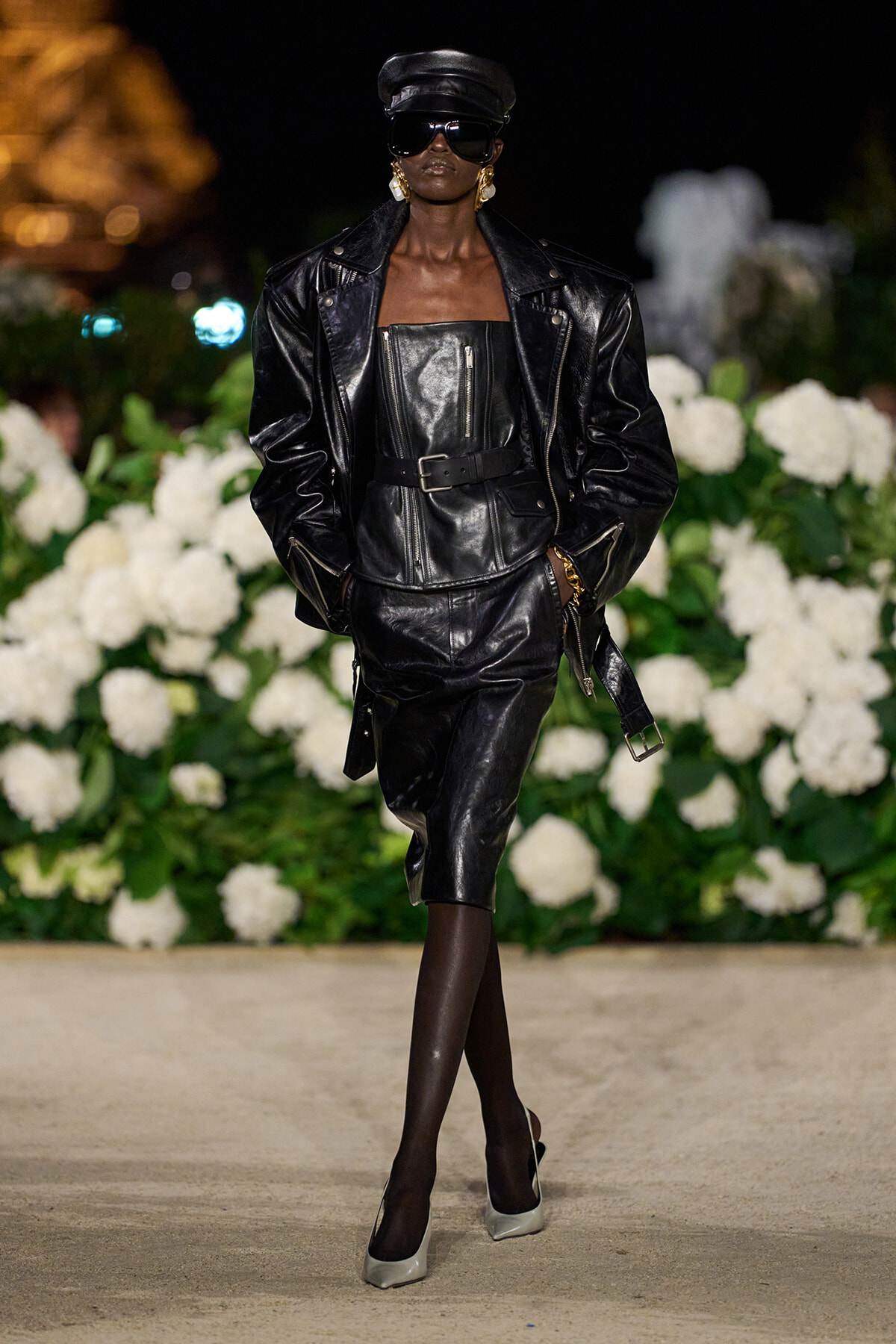 Model in a black leather ensemble (jacket, corset-style top, belt) with a cap and sunglasses walking a runway, white hydrangea backdrop behind.