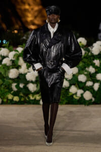 Model in an oversized black leather belted trench, white shirt cuffs, and cap on a runway with white florals in the background.