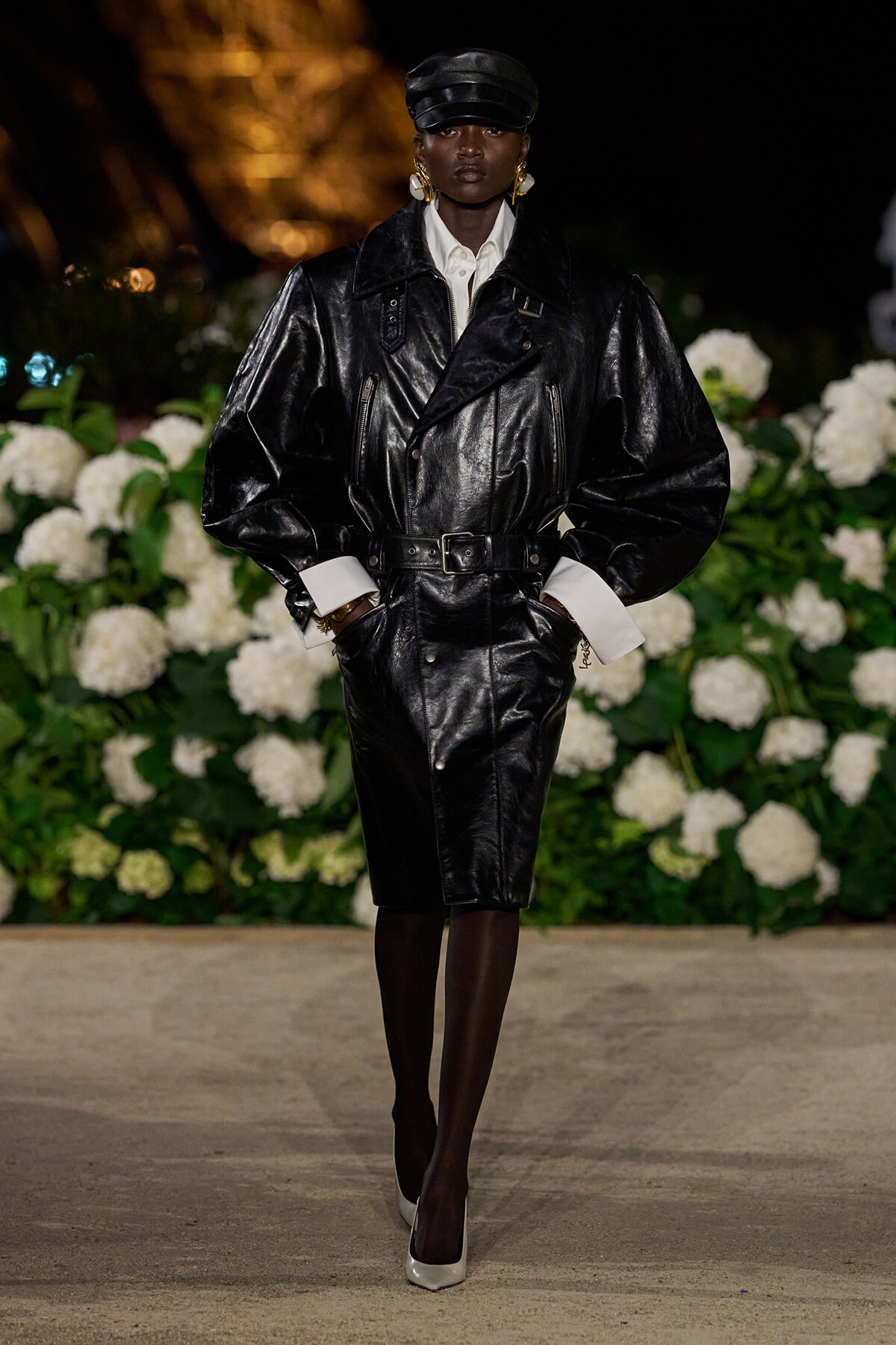 Model in an oversized black leather belted trench, white shirt cuffs, and cap on a runway with white florals in the background.
