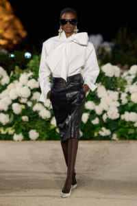 Model on a nighttime runway wearing an oversized white blouse with a large bow and a black leather midi skirt, white heels, and dark sunglasses with statement earrings.