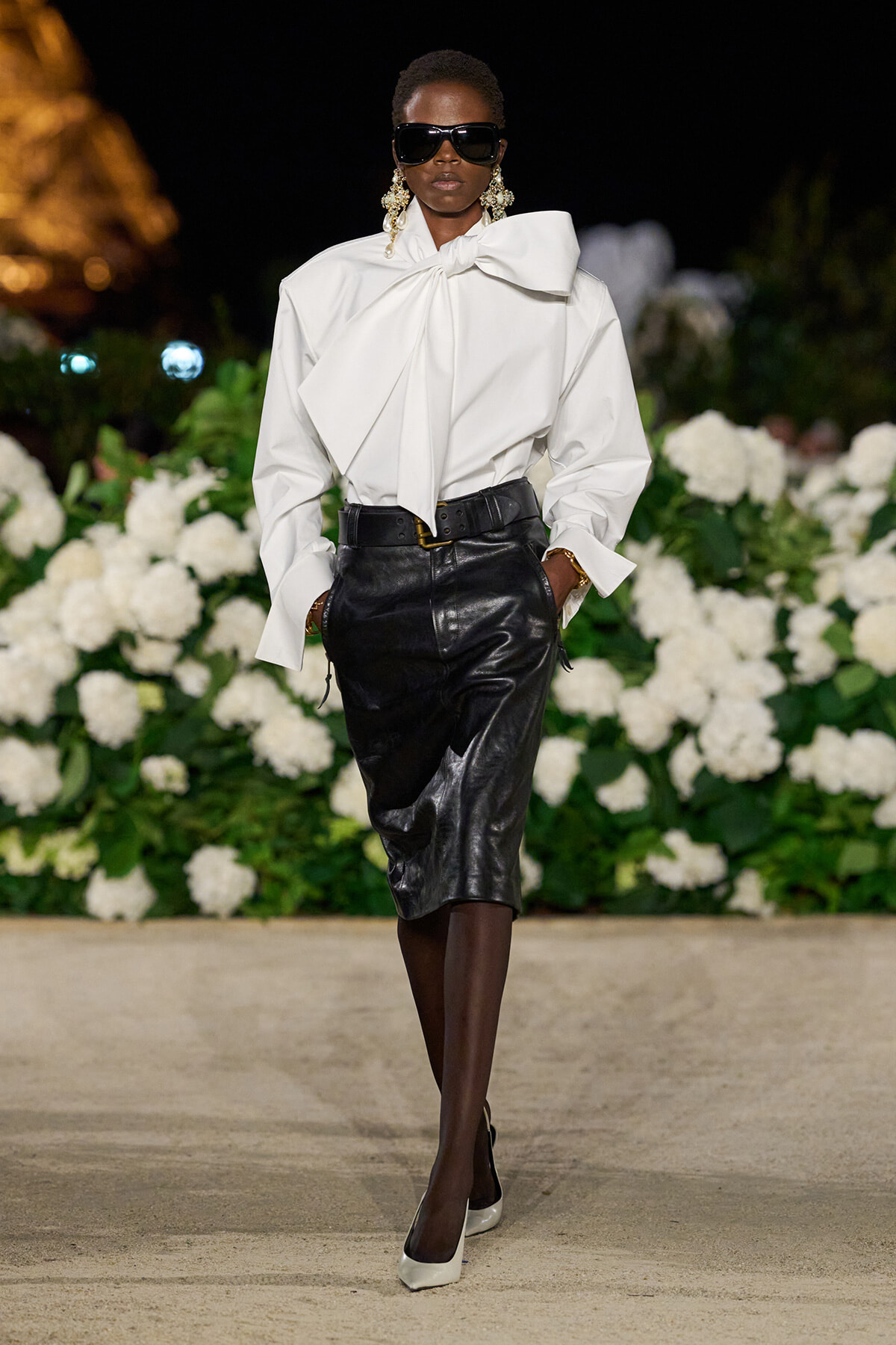 Model on a nighttime runway wearing an oversized white blouse with a large bow and a black leather midi skirt, white heels, and dark sunglasses with statement earrings.