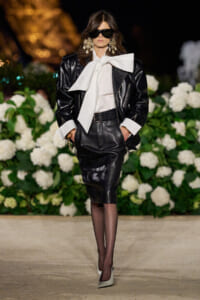 Model walking a runway in a black leather suit with a white oversized bow blouse, statement earrings, and oversized sunglasses, hands in pockets amid white florals backdrop.