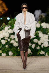 Model wearing a white oversized blouse tied at the waist with a brown leather pencil skirt on a runway, with chunky belt and sunglasses.