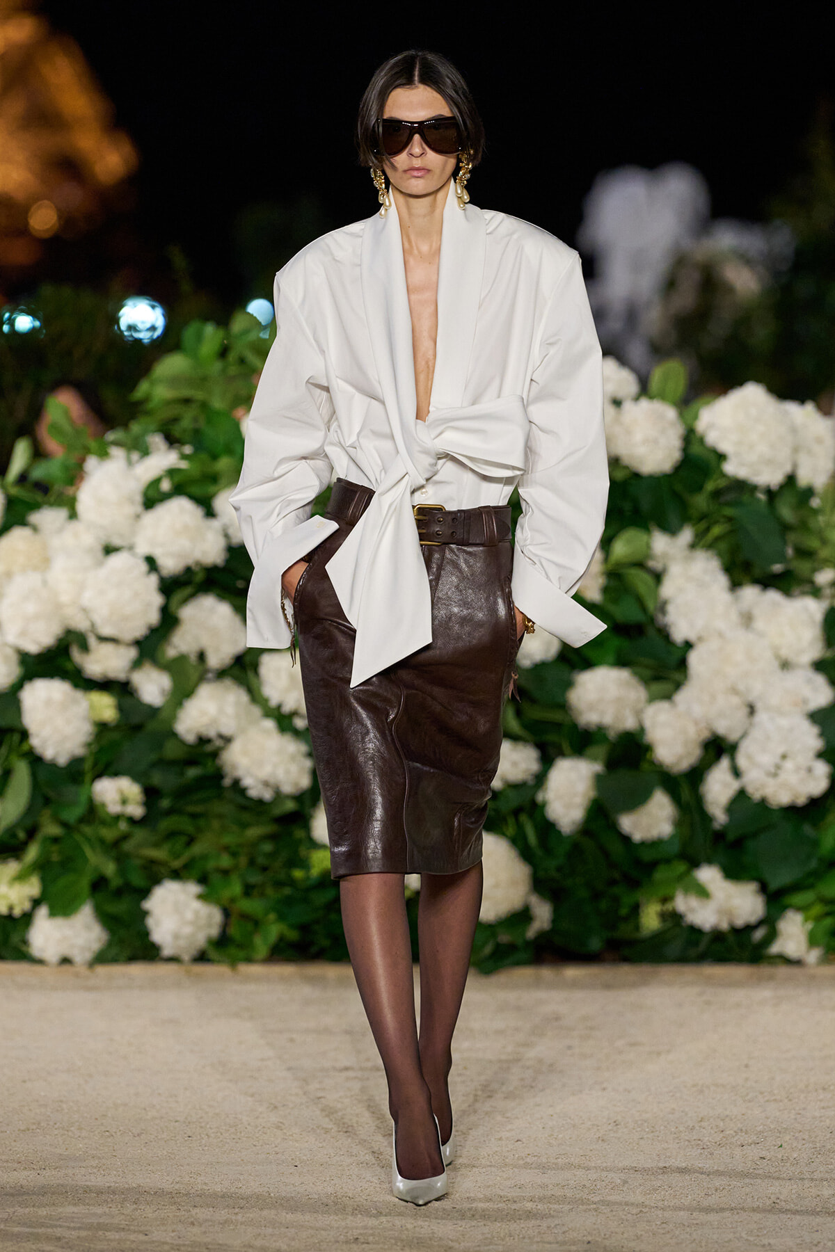 Model wearing a white oversized blouse tied at the waist with a brown leather pencil skirt on a runway, with chunky belt and sunglasses.