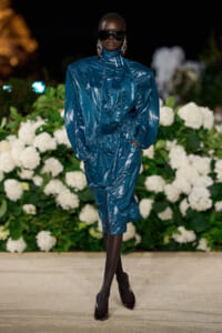 Model walking a nighttime runway in a metallic teal-blue oversized raincoat with a cinched waist, black tights, burgundy heels, and large sunglasses against a backdrop of white hydrangea flowers.