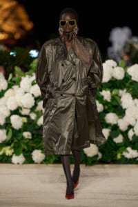 Model wearing an oversized metallic trench coat with yellow sunglasses, bold earrings, and red heels on a floral runway backdrop, at a fashion show.