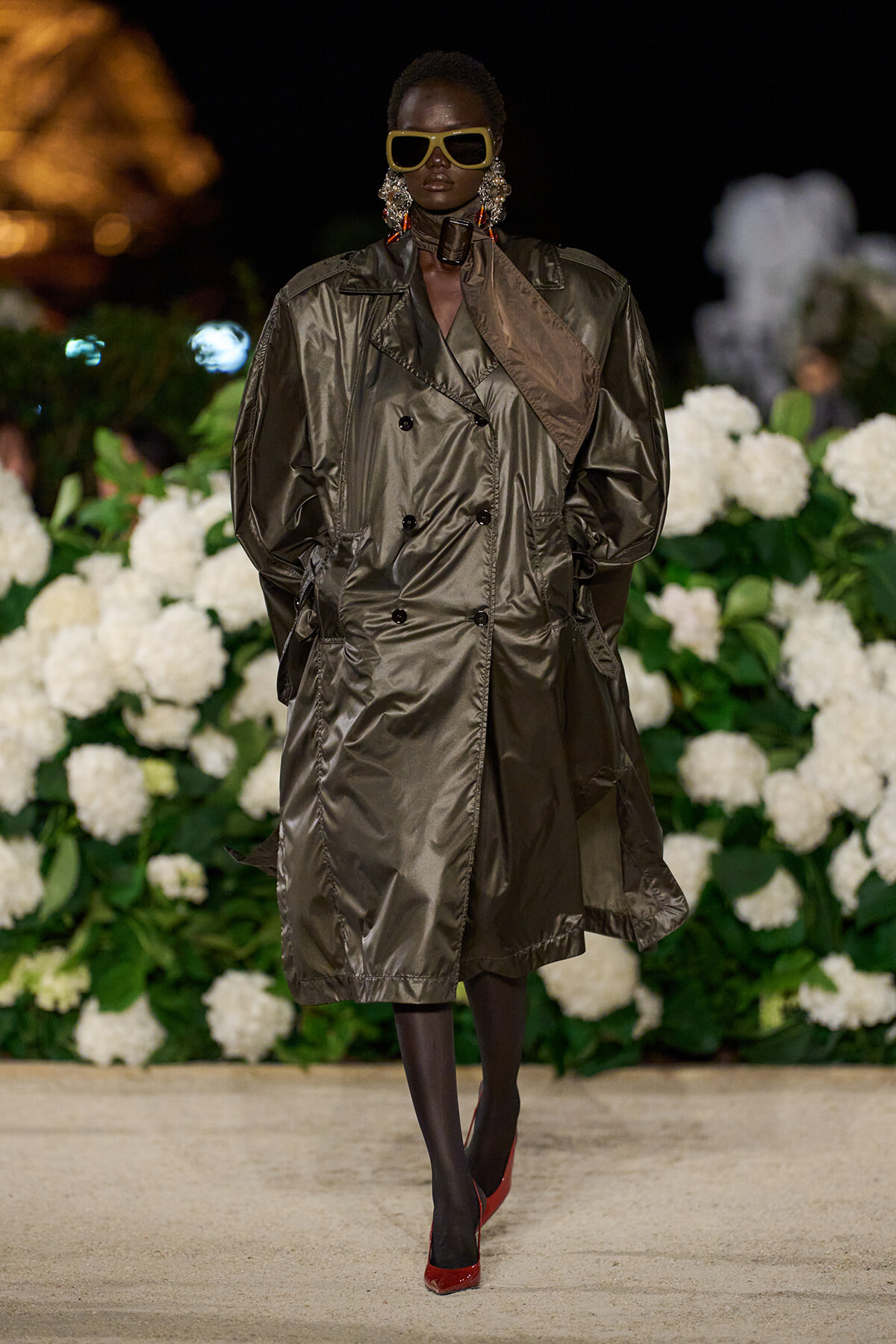 Model wearing an oversized metallic trench coat with yellow sunglasses, bold earrings, and red heels on a floral runway backdrop, at a fashion show.