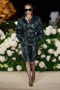 Model on a runway wearing a dark glossy belted coat-dress with oversized sunglasses and bold jewelry, against a backdrop of white hydrangeas.