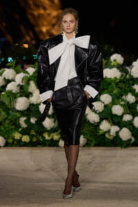 Model walking the runway in a black leather suit with a white oversized bow blouse, gold statement earrings, and gray heels, holding a small black clutch against a backdrop of white hydrangea flowers.