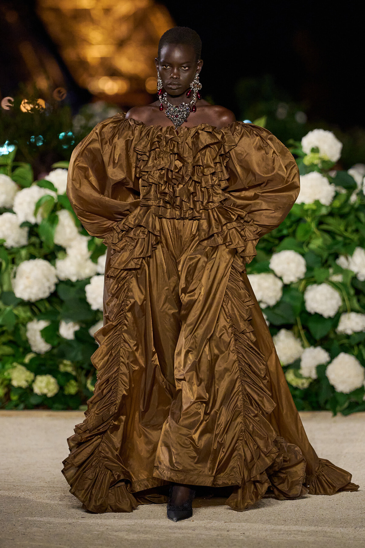 Model on runway wearing a metallic bronze off-shoulder ballgown with tiered ruffles and voluminous sleeves, accessorized with bold jewelry, floral backdrop