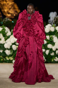 Model in a dramatic burgundy gown with oversized, ruffled sleeves on a nighttime runway, with white flowers in the background.