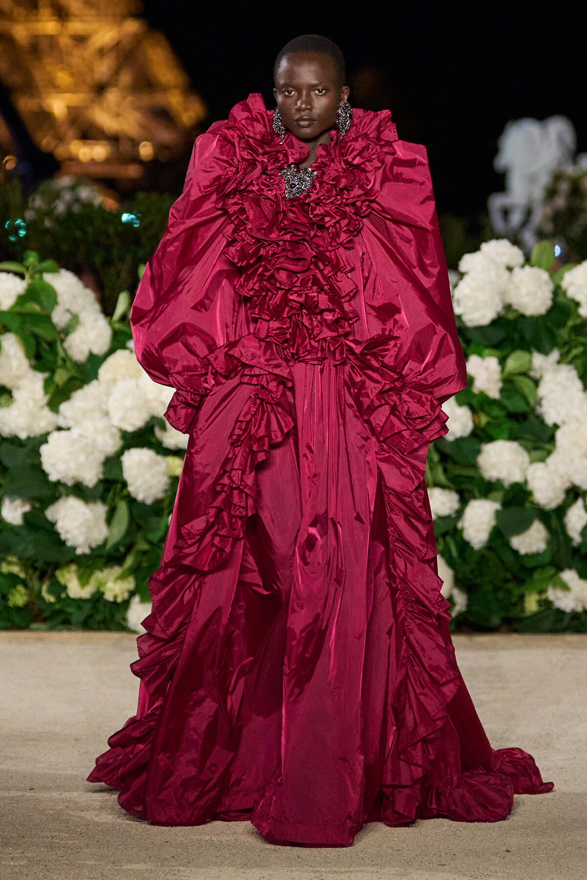 Model in a dramatic burgundy gown with oversized, ruffled sleeves on a nighttime runway, with white flowers in the background.