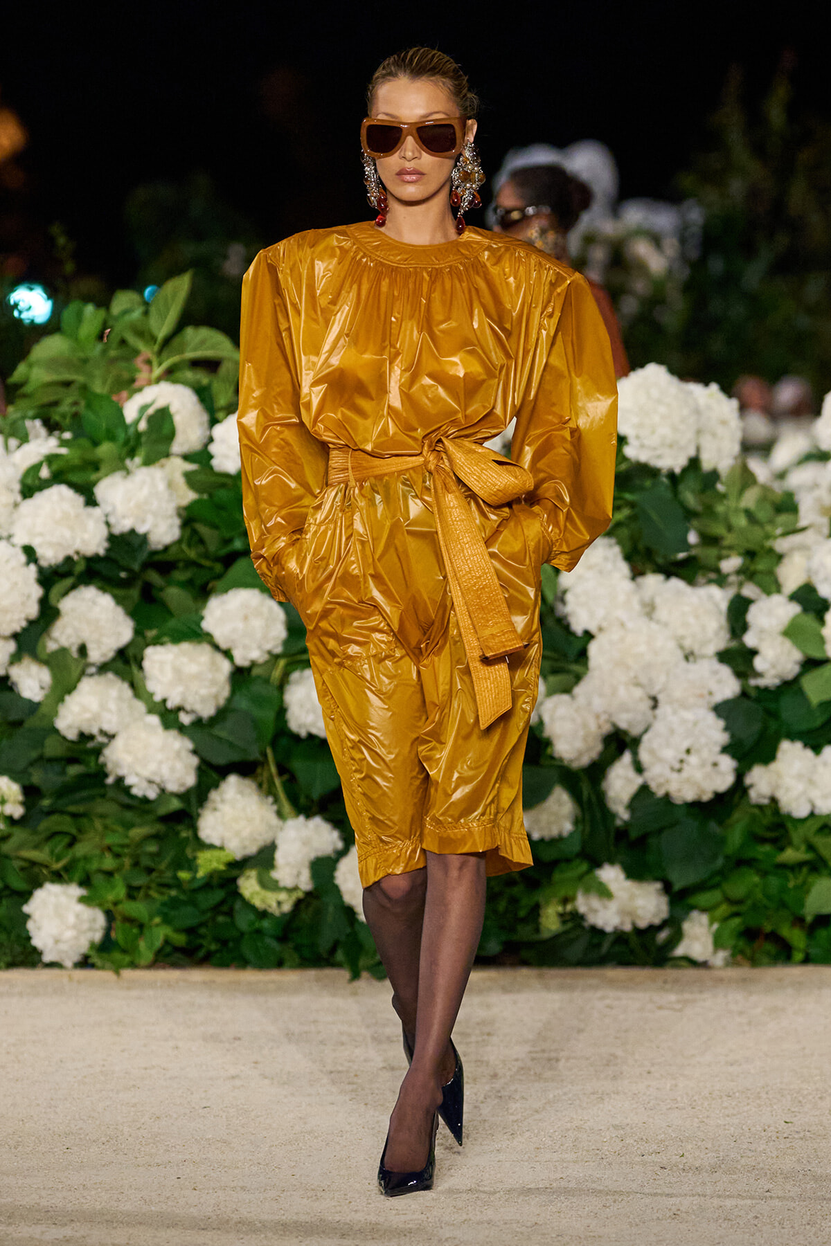 Model in a glossy mustard-yellow jumpsuit with a wide belt, wearing oversized sunglasses and statement earrings on a floral runway backdrop.