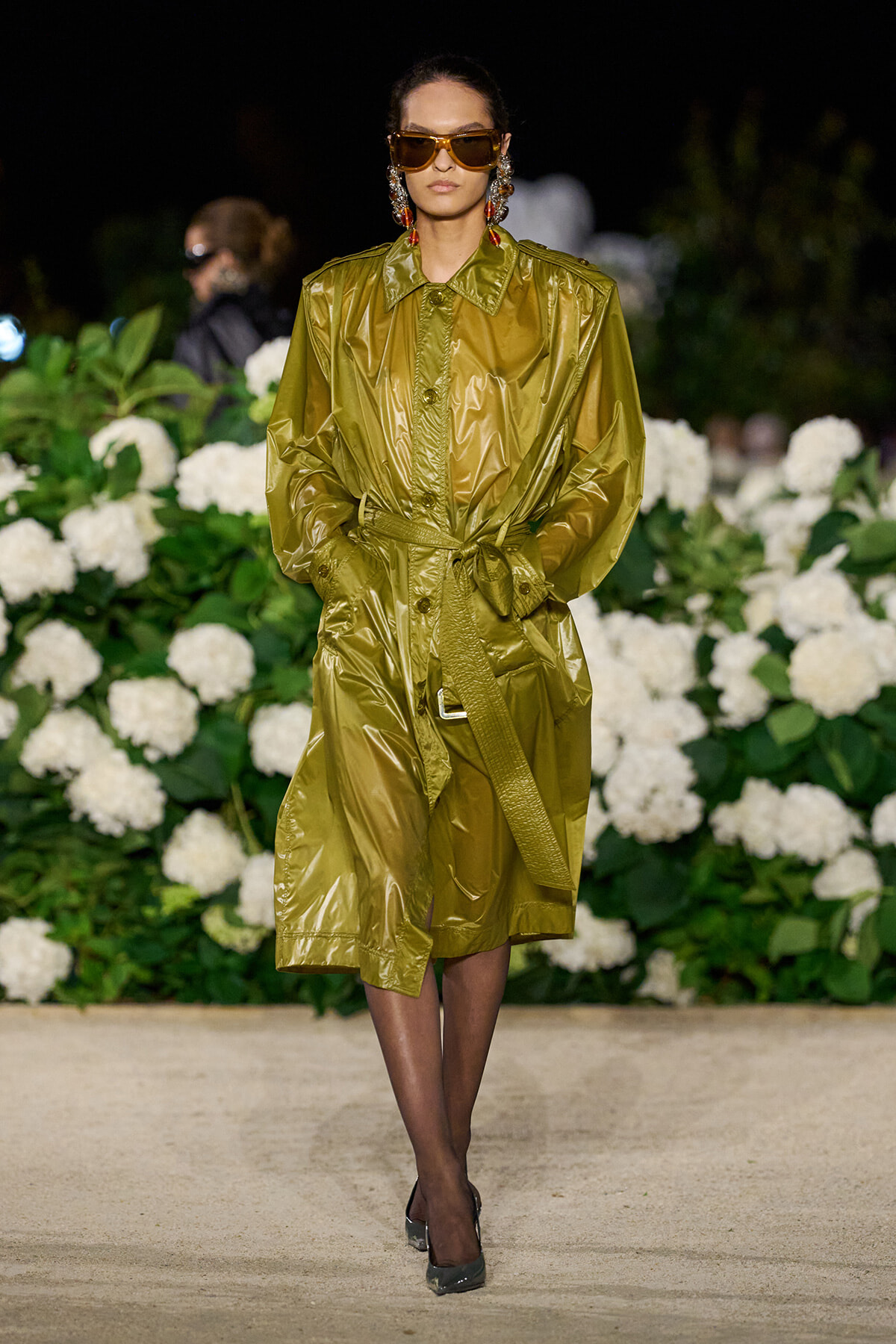 Model in a metallic olive trench coat with a tied waist, oversized sunglasses, and bold earrings on a floral runway backdrop.