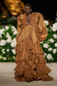 Model walking a nighttime runway in a bronze-brown, tiered ruffled gown with dramatic puff sleeves and ornate jewelry.