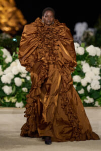 Model wearing a dramatic bronze-brown, heavily ruffled gown on a runway with white hydrangeas in the background.