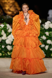 Model walking a runway in a vivid orange, multi-tiered ruffled ball gown with dramatic puff sleeves and a statement necklace, floral backdrop behind her.