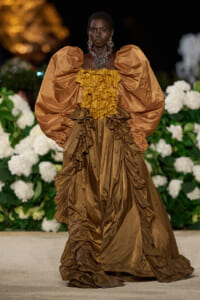 Model on a runway wearing a bronze-brown gown with a gold ruffled bodice and oversized puff sleeves.