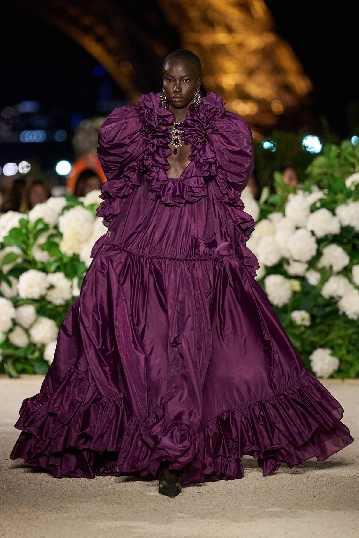 Model wearing a deep purple, floor-length ruffled gown with oversized shoulders walking a runway at night among white flower arrangements.
