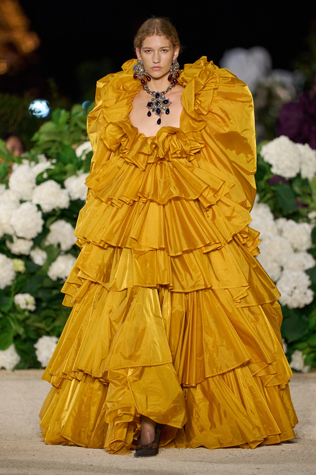 Model on a runway wearing a voluminous mustard-yellow ruffled gown with bold jewelry, standing among white hydrangeas at night.