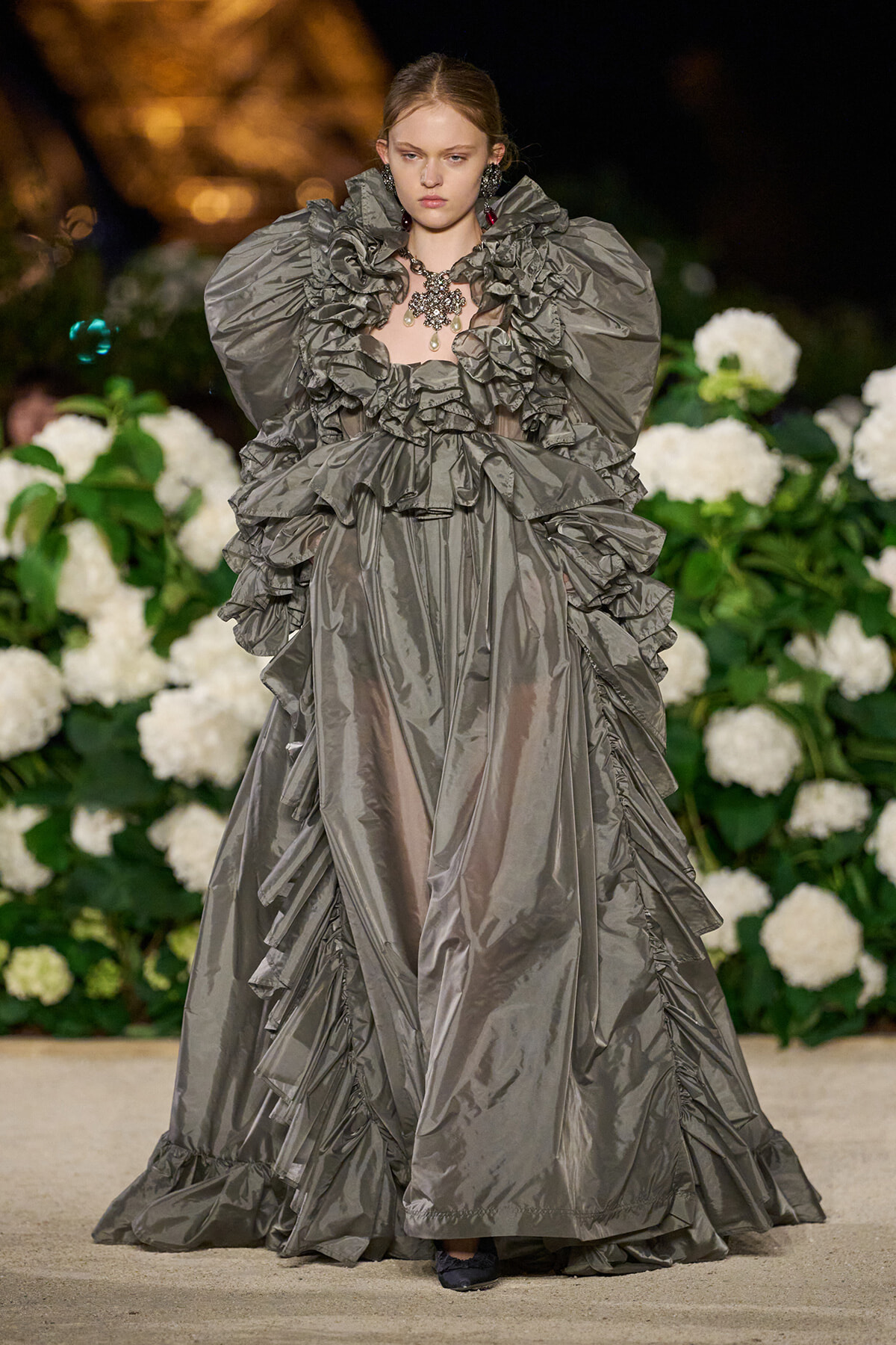 Model wearing a dramatic metallic gray gown with oversized ruffles on a runway, against a backdrop of white flowers and greenery.