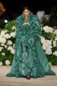Model on a runway wearing an emerald green gown with oversized puff sleeves and cascading ruffles, paired with bold jewelry, against a backdrop of white hydrangeas.