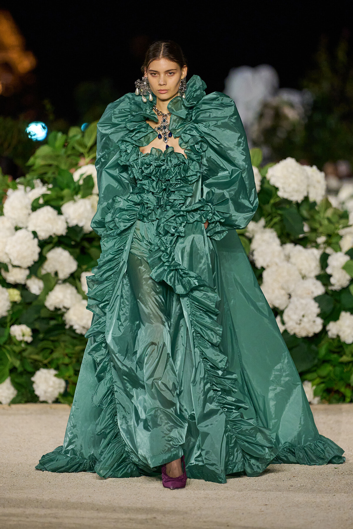 Model on a runway wearing an emerald green gown with oversized puff sleeves and cascading ruffles, paired with bold jewelry, against a backdrop of white hydrangeas.