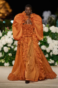 Model walking a runway in an orange, voluminous, ruffled gown with dramatic puff sleeves against a backdrop of white hydrangeas at night