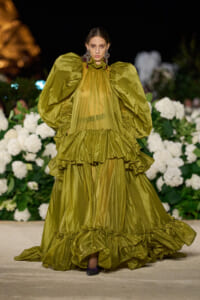 Model walks the runway in an olive-green, voluminous gown with oversized puff sleeves and layered ruffles, backdrop of white flowers.