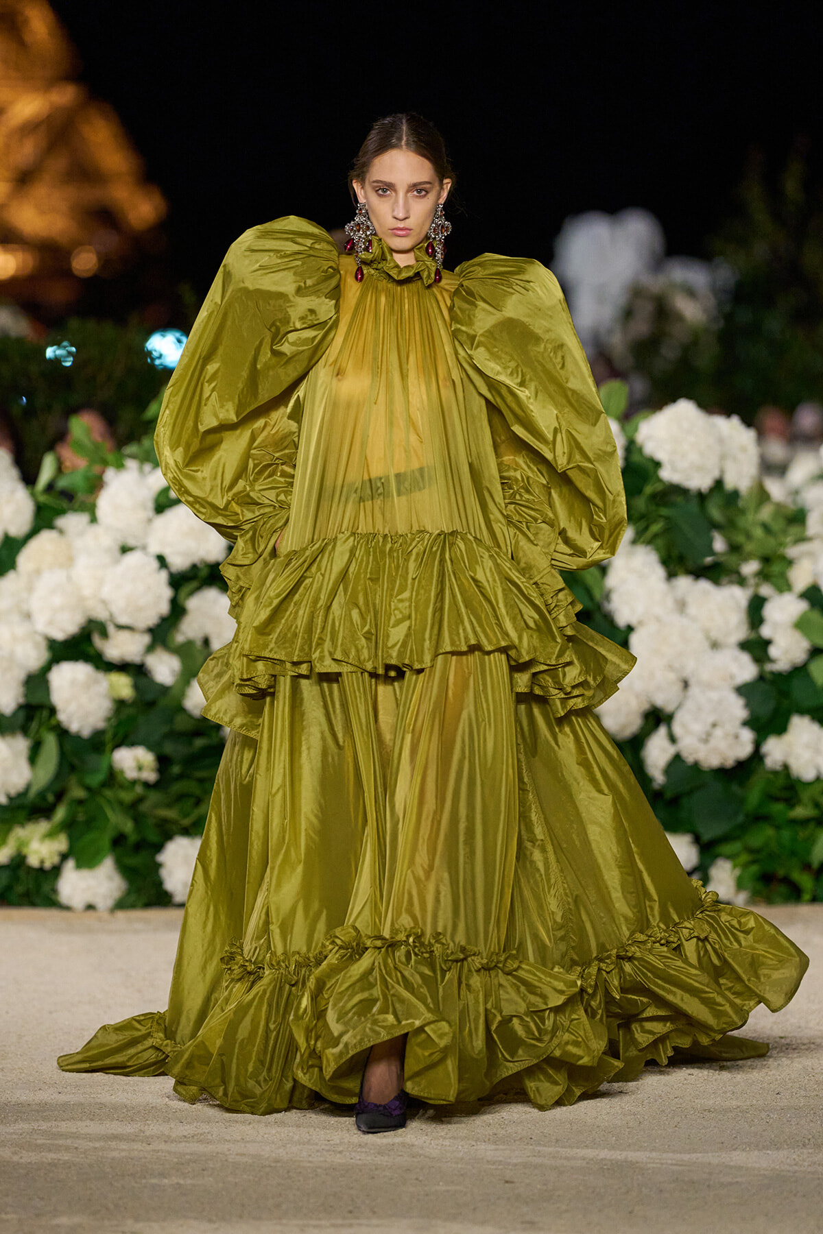 Model walks the runway in an olive-green, voluminous gown with oversized puff sleeves and layered ruffles, backdrop of white flowers.