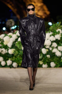 Model on a runway wearing a long black shiny vinyl trench with a waist tie, oversized shoulders, and black sunglasses against a backdrop of white hydrangeas.