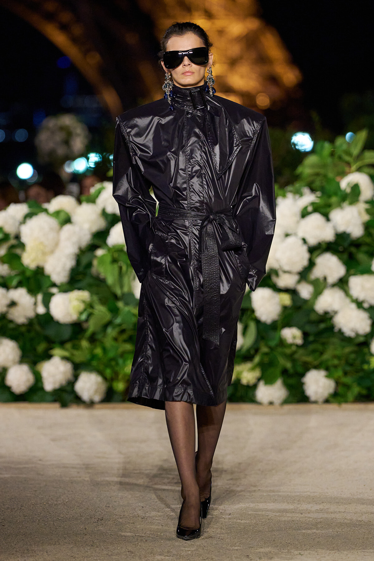 Model on a runway wearing a long black shiny vinyl trench with a waist tie, oversized shoulders, and black sunglasses against a backdrop of white hydrangeas.