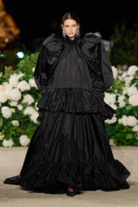 Runway model wearing a dramatic black gown with oversized puff sleeves and multiple tiered ruffles, standing in front of white floral backdrop at night.