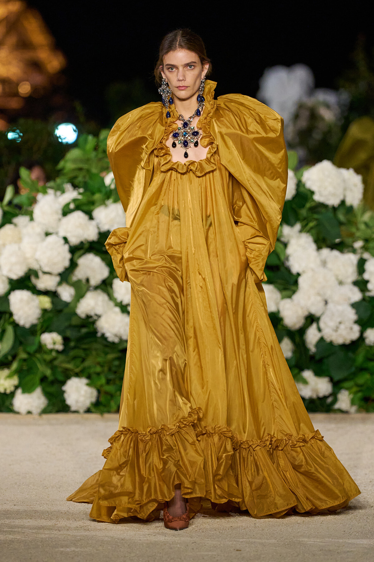Model on a runway wearing a gold satin gown with oversized puff sleeves and a ruffled hem, accessorized with a blue jeweled necklace against a floral backdrop.