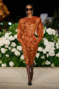 Model walking the runway in a metallic orange belted trench dress with oversized sleeves, dark sunglasses, and statement earrings, against a floral backdrop.