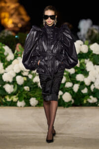 Model walking a runway in a glossy black puffer with oversized sleeves, cinched waist, black heels, and large sunglasses against a floral backdrop.