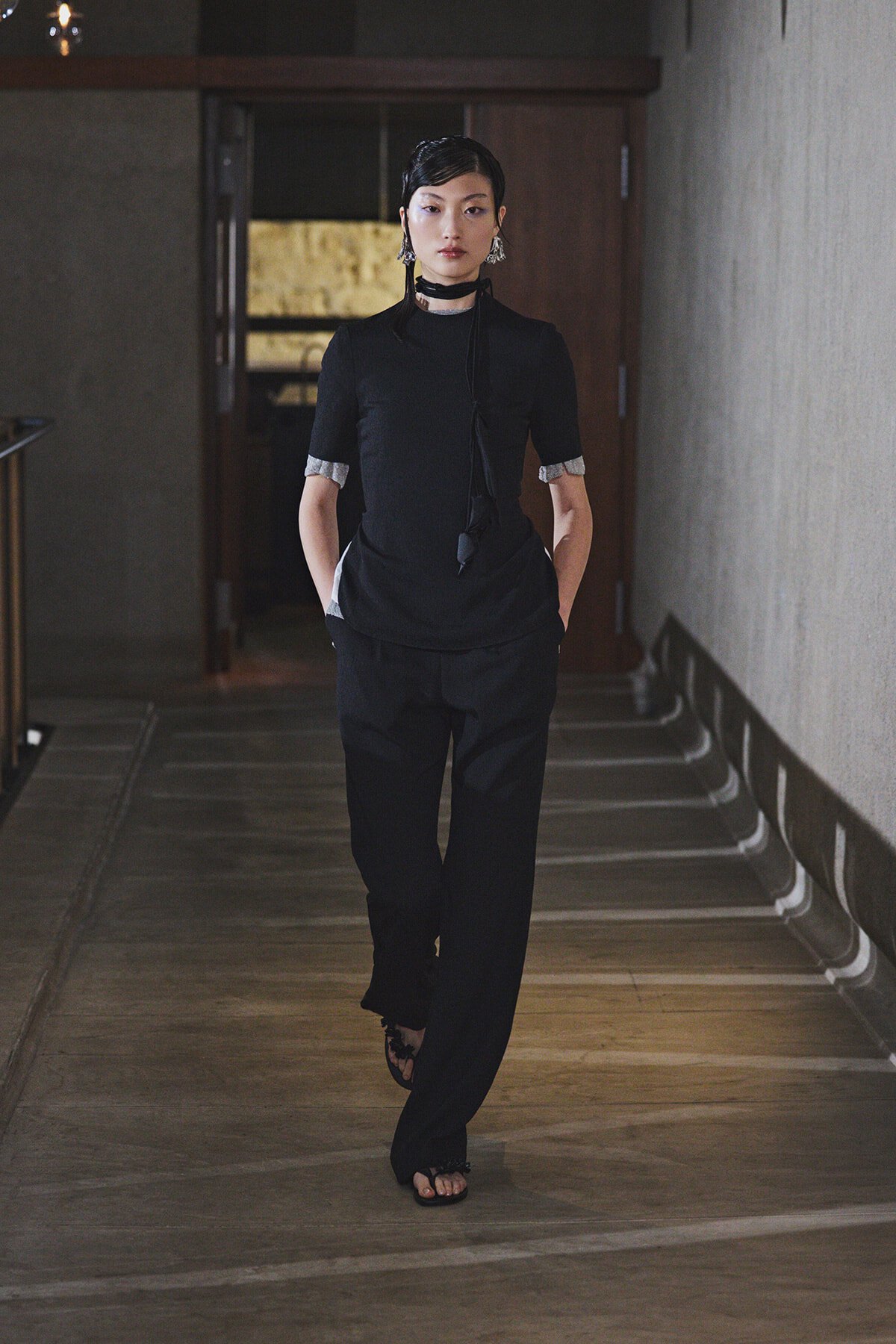 Woman in an all-black ensemble walking down a dim hallway with hands in pockets, wearing a high-neck top, scarf, choker, and sandals.