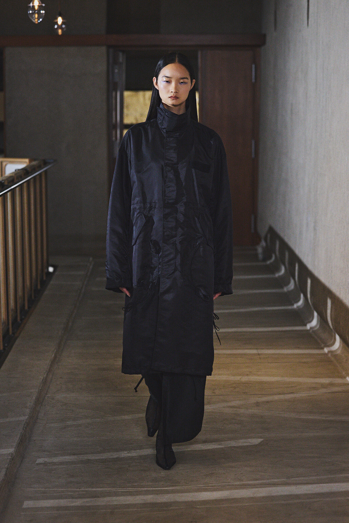 Young woman in a long black coat walking toward camera in a dim indoor hallway.