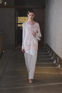 Model walking in a dim hallway wearing a pale pink sheer top, white textured pants, and a small transparent handbag.