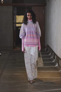Woman with curly hair wearing a pastel pink-lilac oversized sweater and floral white skirt, carrying a woven beige bag in a dim hallway.