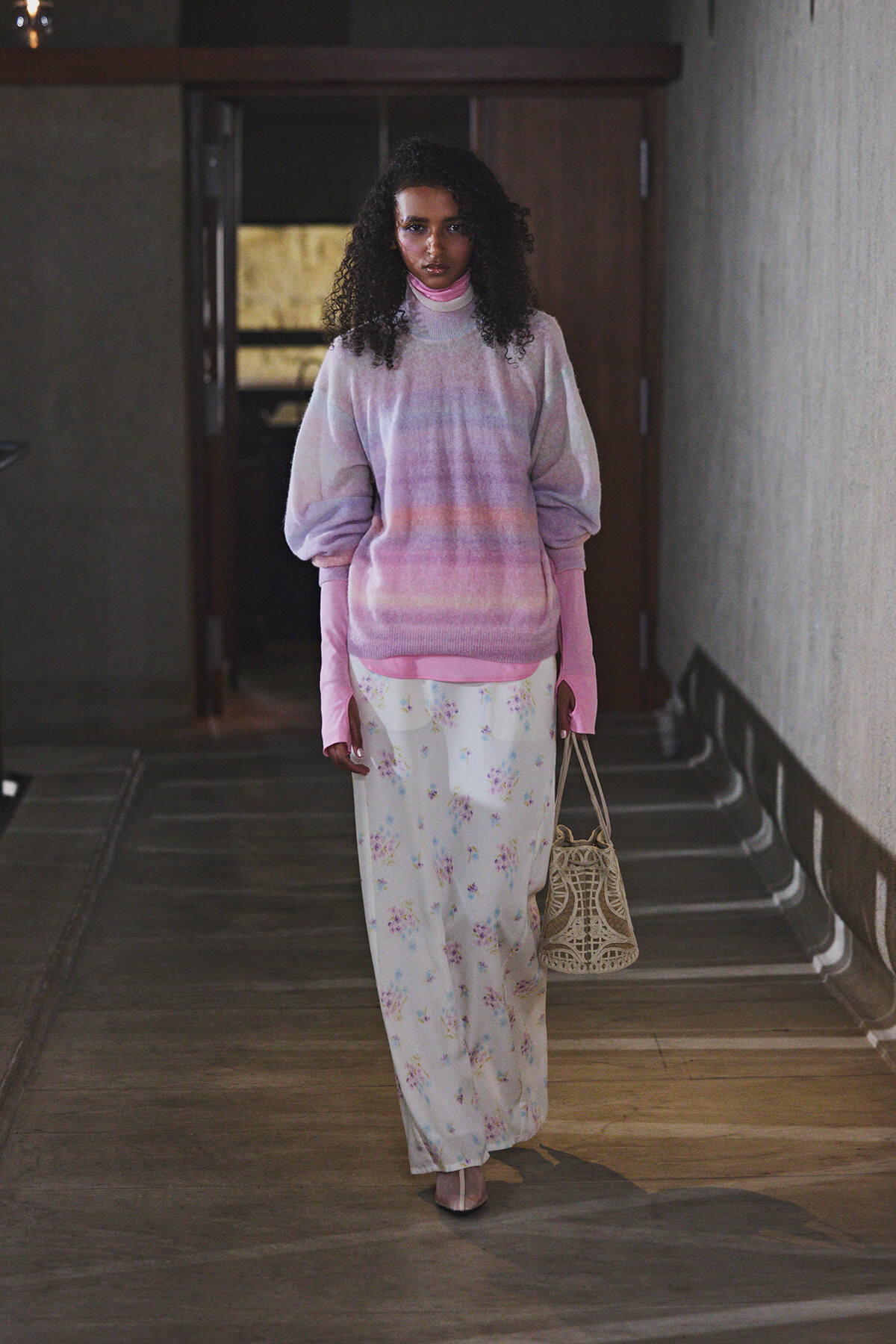Woman with curly hair wearing a pastel pink-lilac oversized sweater and floral white skirt, carrying a woven beige bag in a dim hallway.