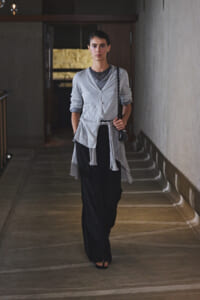 Model walking toward the camera in a dim hallway, wearing a light gray cardigan over a gray top with black wide-leg pants and a chain-strap bag on her shoulder.