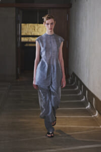 Model walking down a dim hallway wearing a light gray sleeveless tunic and loose matching pants in a minimalist fashion look.