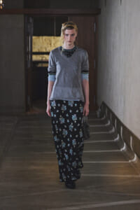 Woman walking down a dim hallway in a gray V-neck top over a black floral maxi skirt, carrying a small bag.